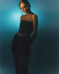 Portrait of a woman wearing a dark strapless top and long earrings against a gradient blue and warm-lit background.