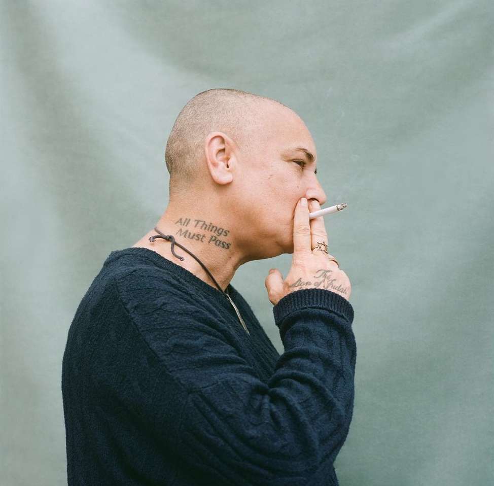 Profile shot of an adult smoking a cigarette. The person has a shaved head, neck tattoo, and wears a dark sweater.