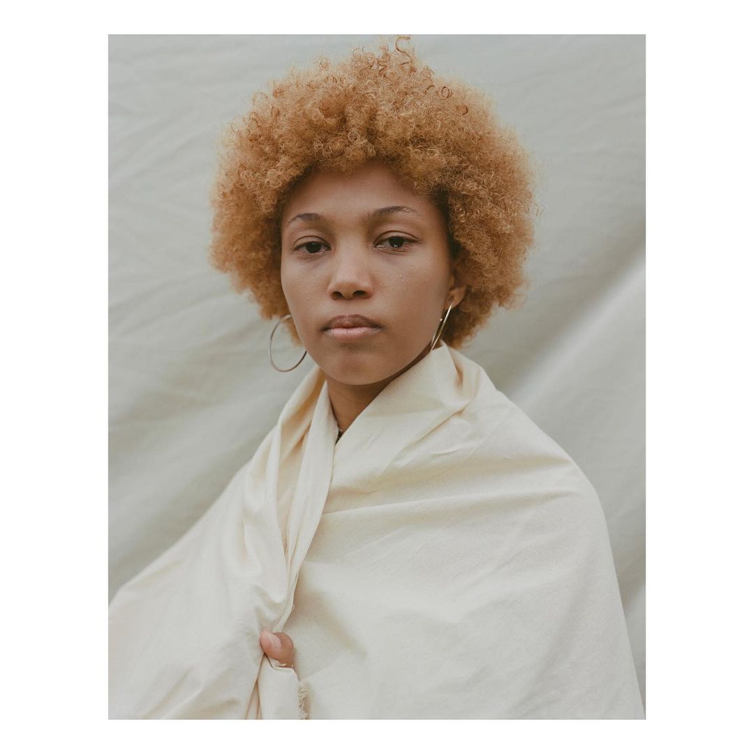 Portrait of a young adult female with curly blonde hair wrapped in a cream-colored fabric against a muted background in soft, diffused light.