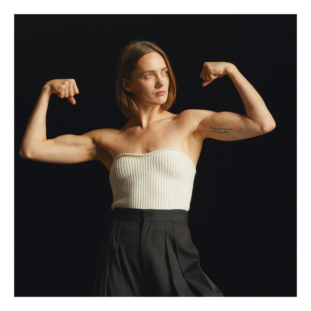 Fit adult female flexing biceps in a studio environment. She is wearing a strapless ribbed top and pleated pants against a black backdrop.