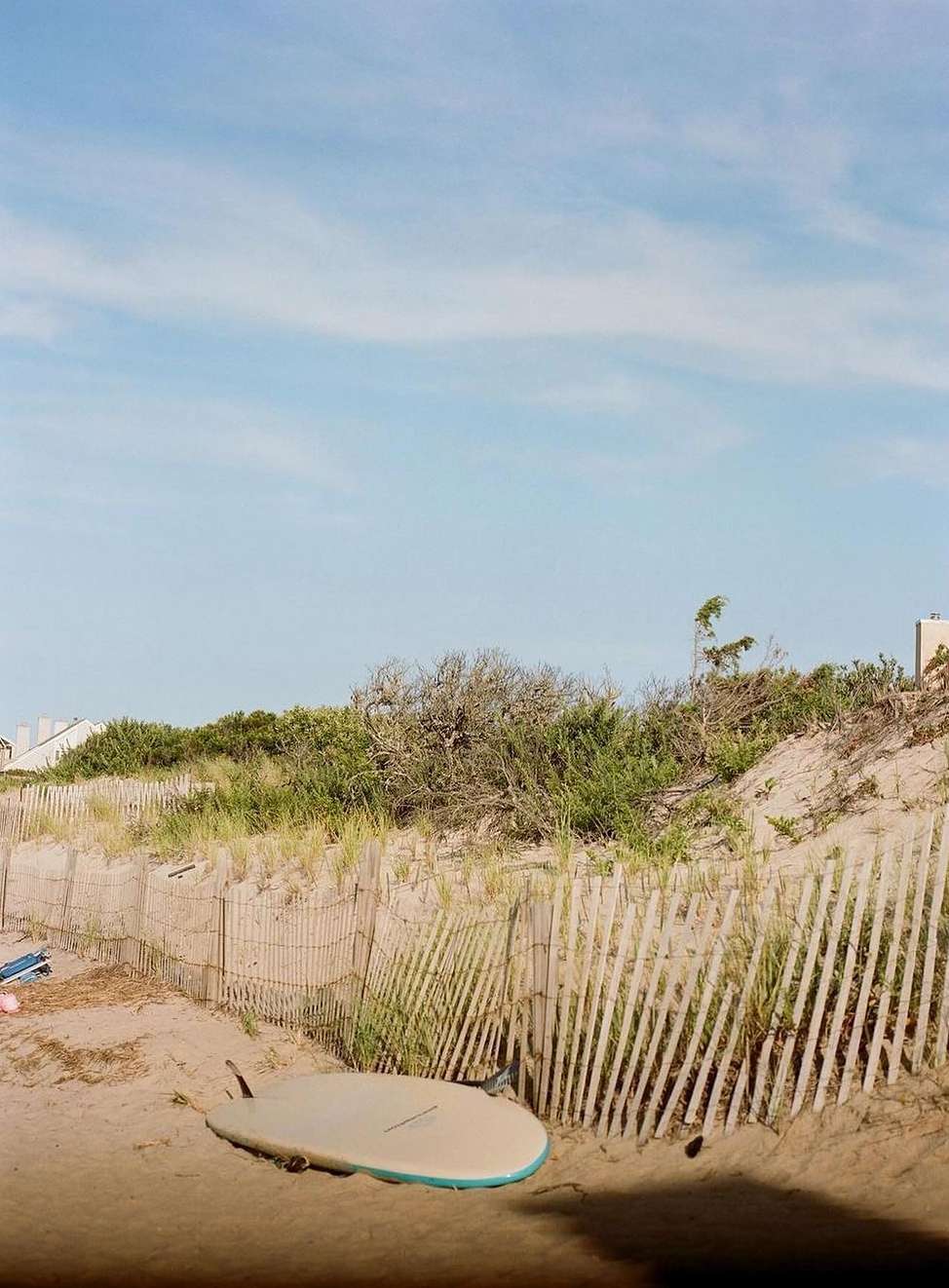 Surfboard rests on a sandy beach next to a wooden fence under a sunny, blue sky with light clouds on a bright sunny day.