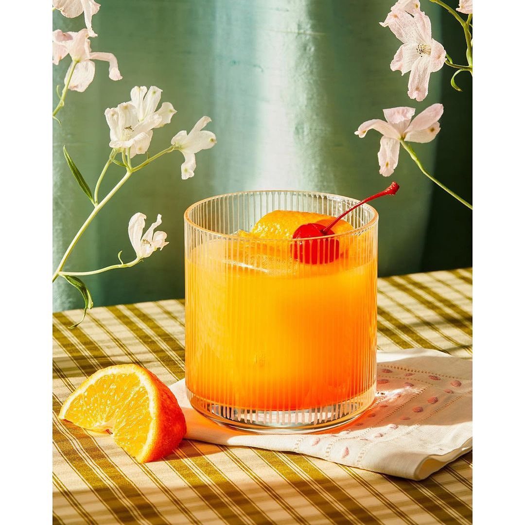 Refreshing drink in ribbed glass with orange and cherry garnish. Beverage on checkered table with white blossoms and green backdrop.