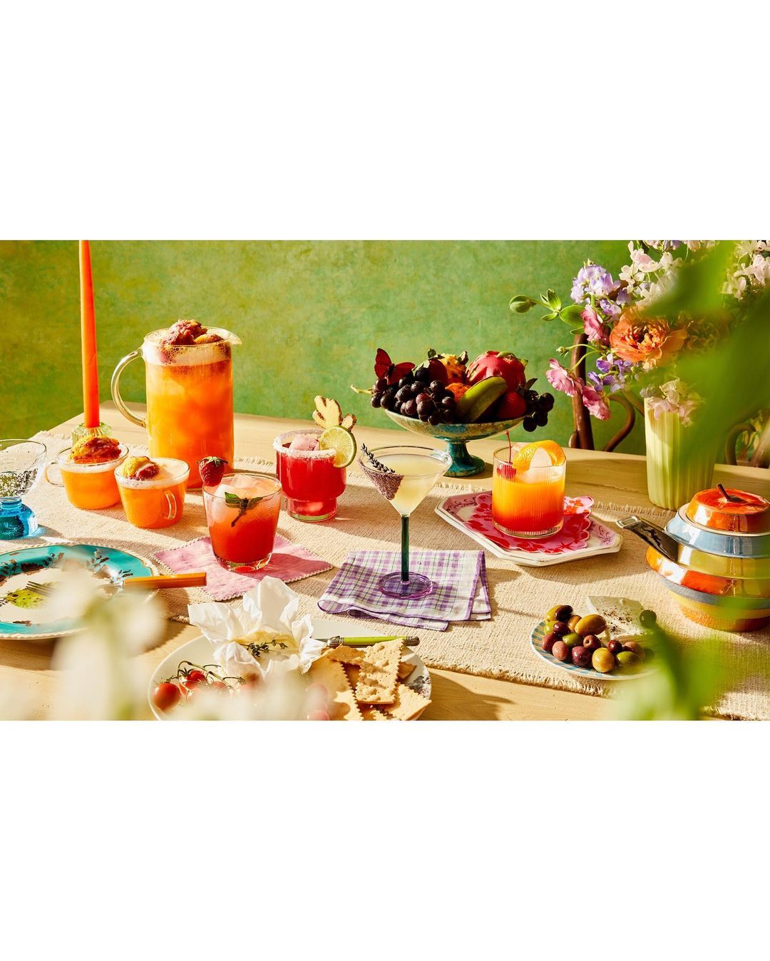 A colorful table spread featuring various cocktails in glassware, a fruit bowl, flowers, and appetizers arranged on a textured cloth.