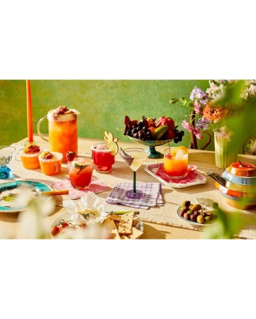 A colorful table spread featuring various cocktails in glassware, a fruit bowl, flowers, and appetizers arranged on a textured cloth.