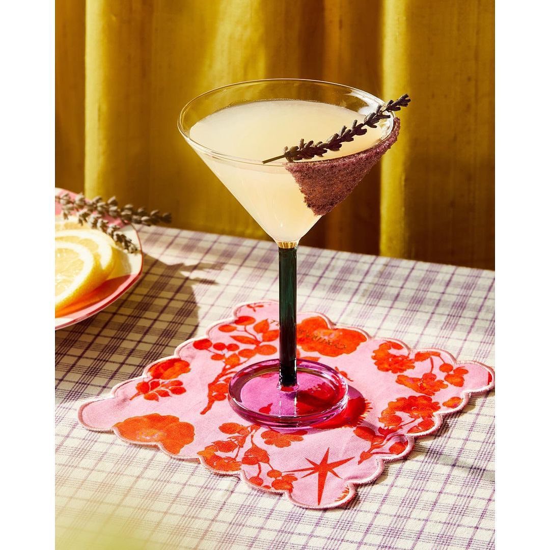Cocktail in a martini glass with a sugared rim and lavender sprig, set on a pink floral coaster on a checkered tablecloth.