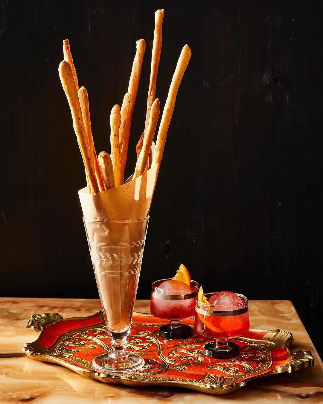Grissini breadsticks in a glass next to two cocktails with orange slices on an ornate tray, set against a dark background.