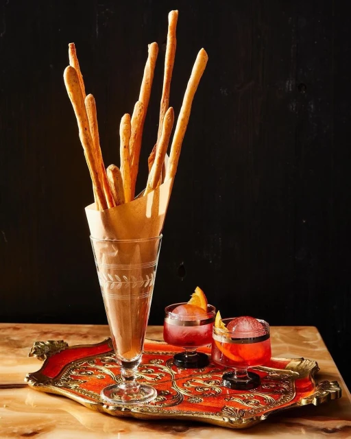Grissini breadsticks in a glass next to two cocktails with orange slices on an ornate tray, set against a dark background.