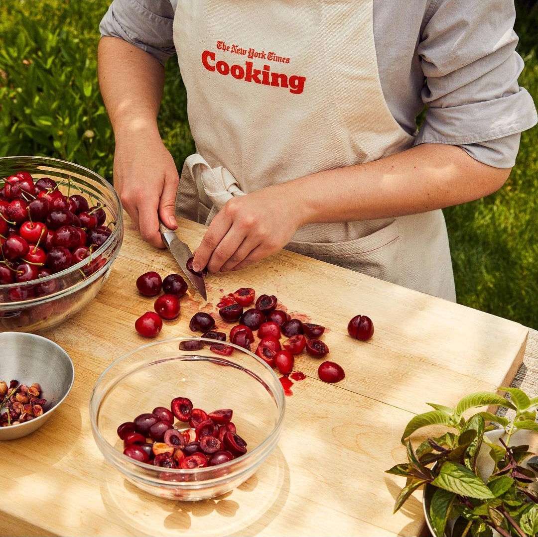 Person in apron prepping red cherries with knife on wooden board, surrounded by glass bowls outdoors in daytime. Food prep concept.