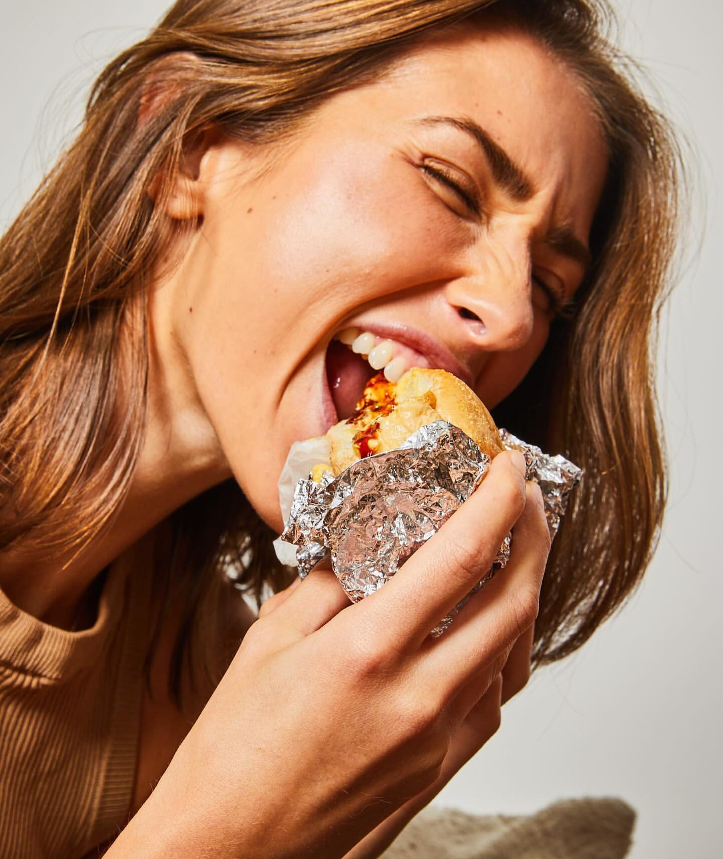 A woman with long brown hair takes a big bite of a sandwich wrapped in foil, showcasing a close-up of the food and her expression.