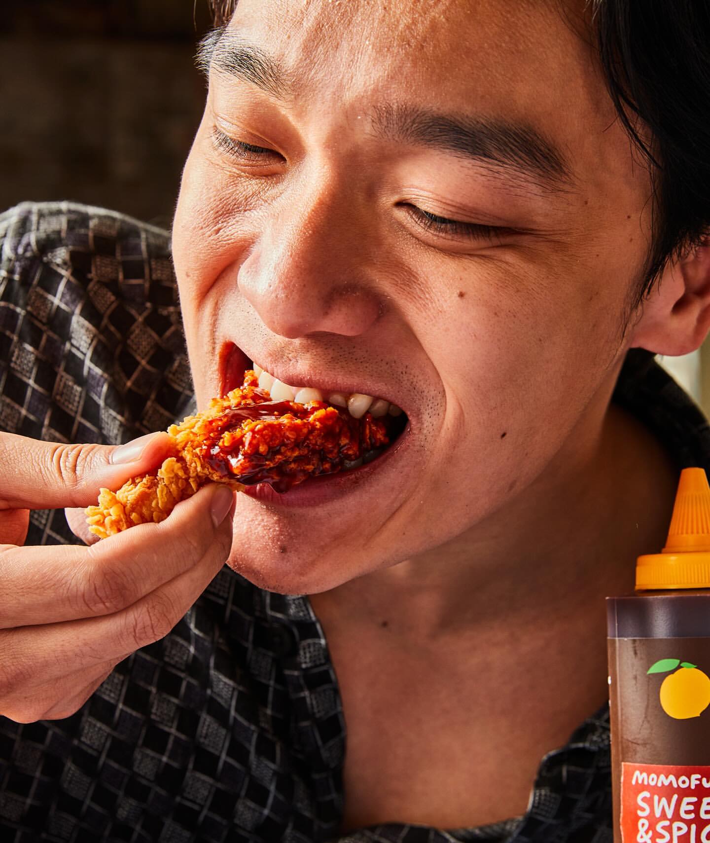Close-up of a person eating a piece of fried chicken covered in sauce indoors, with a bottle of sweet & spicy sauce visible.