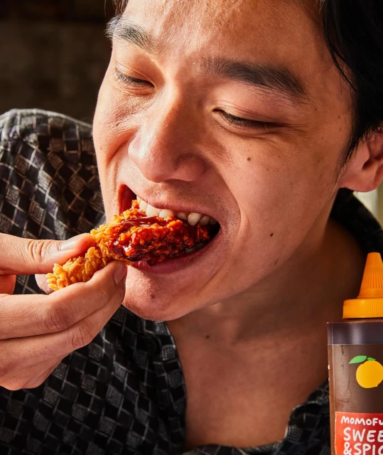 Close-up of a person eating a piece of fried chicken covered in sauce indoors, with a bottle of sweet & spicy sauce visible.