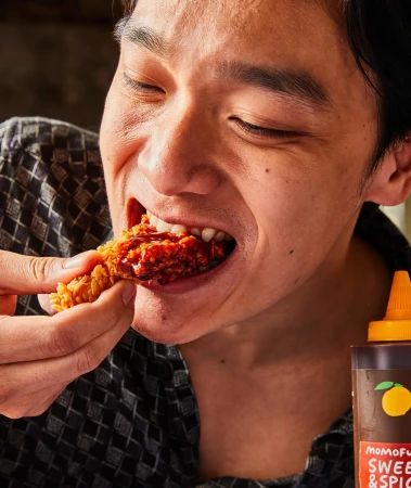 Close-up of a person eating a piece of fried chicken covered in sauce indoors, with a bottle of sweet & spicy sauce visible.