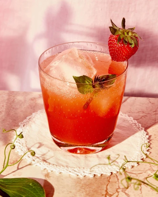 A refreshing strawberry cocktail with ice, garnished with fresh basil and a strawberry, presented on a decorative doily and marble surface.