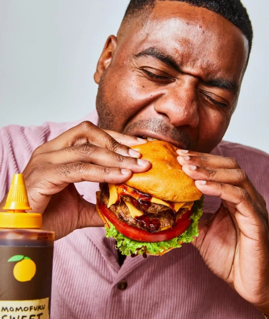 African-American man eating double cheeseburger with tomato, lettuce, and sauce with bottle of Momofuku Sweet & Spicy seasoning.