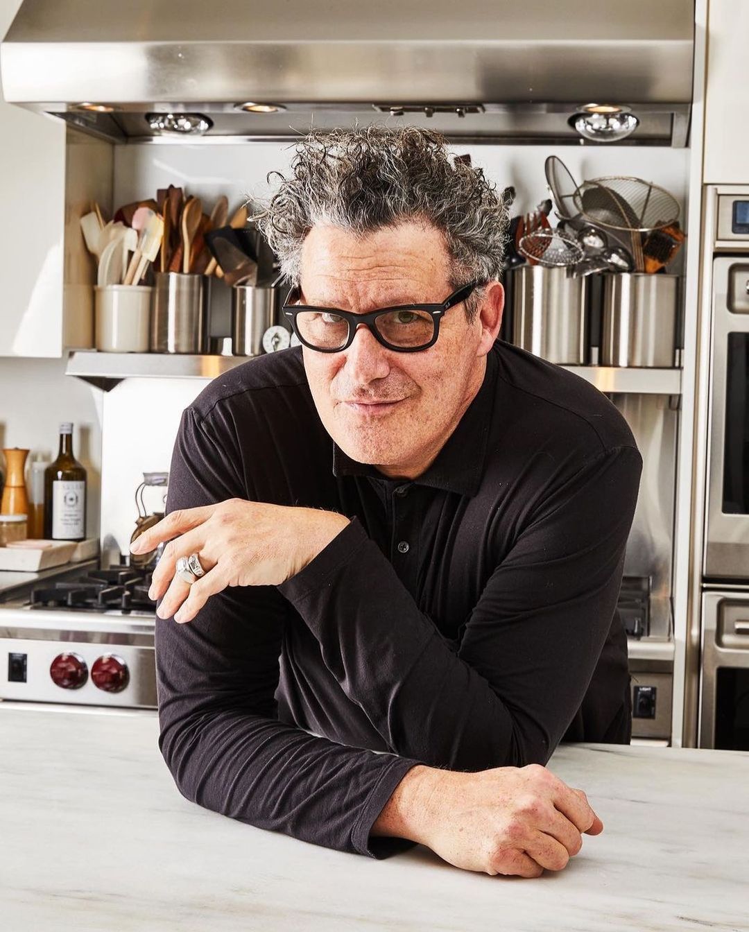 Portrait of adult male with glasses leaning on marble kitchen counter, wearing a black long-sleeve shirt, with stainless steel appliances in background.