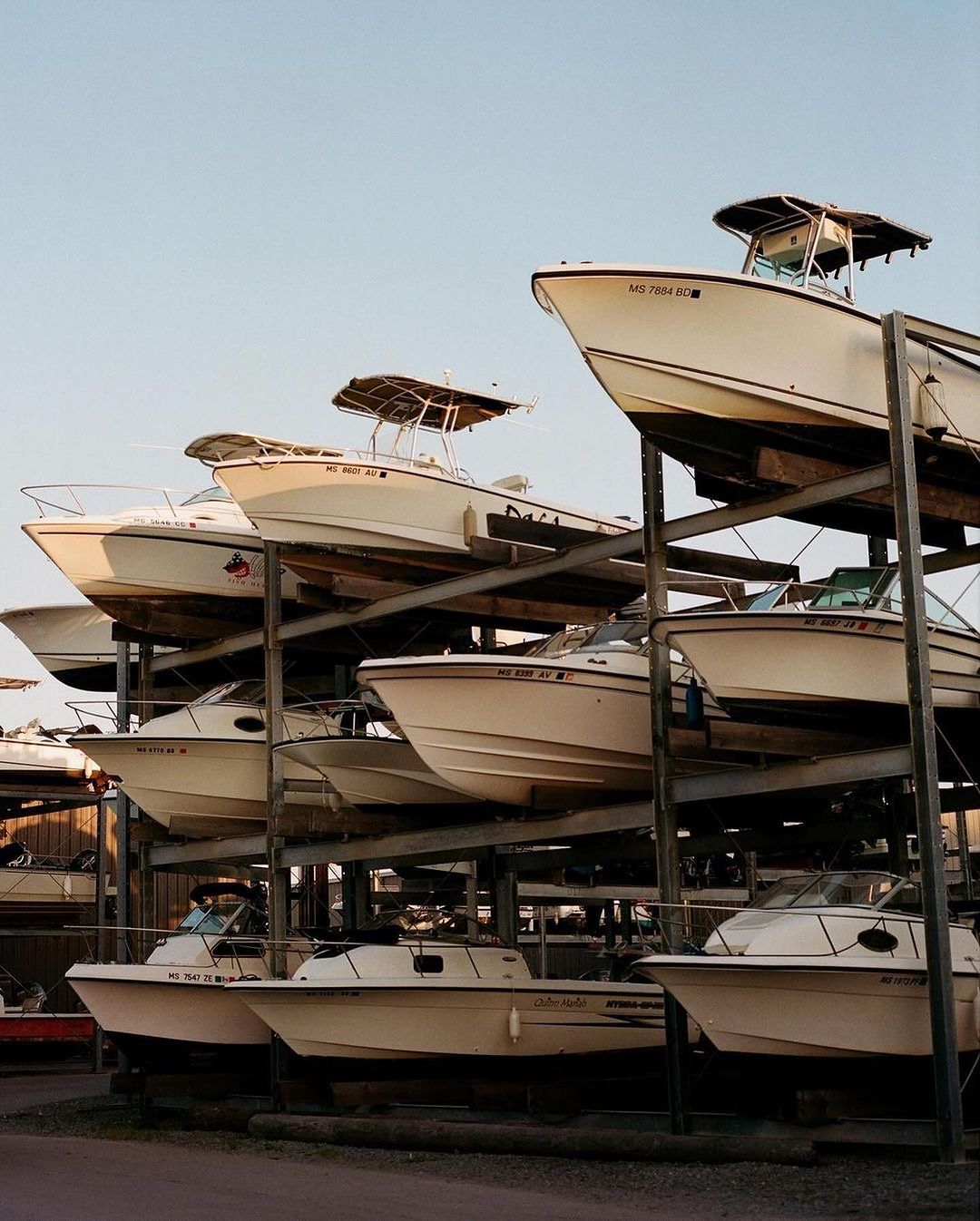 Several cream-colored motorboats are stacked on a metal rack system on a gravel lot under a clear blue sky. Transportation theme.