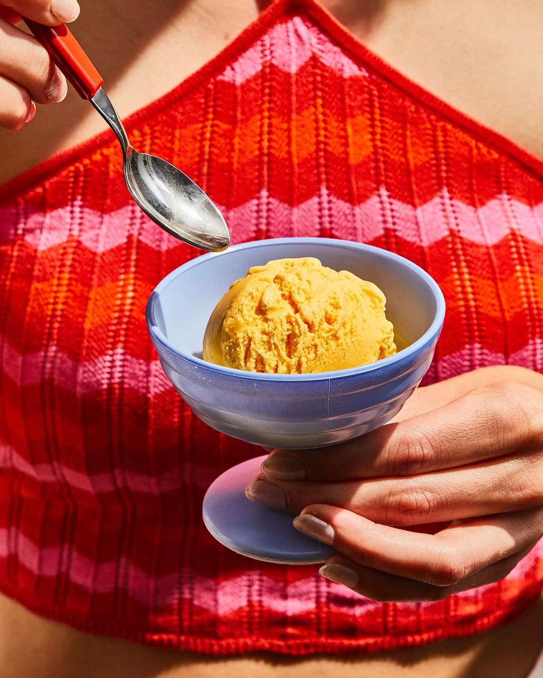 Person holding a bowl of yellow ice cream with a spoon about to scoop some up, against a red and orange chevron patterned top.