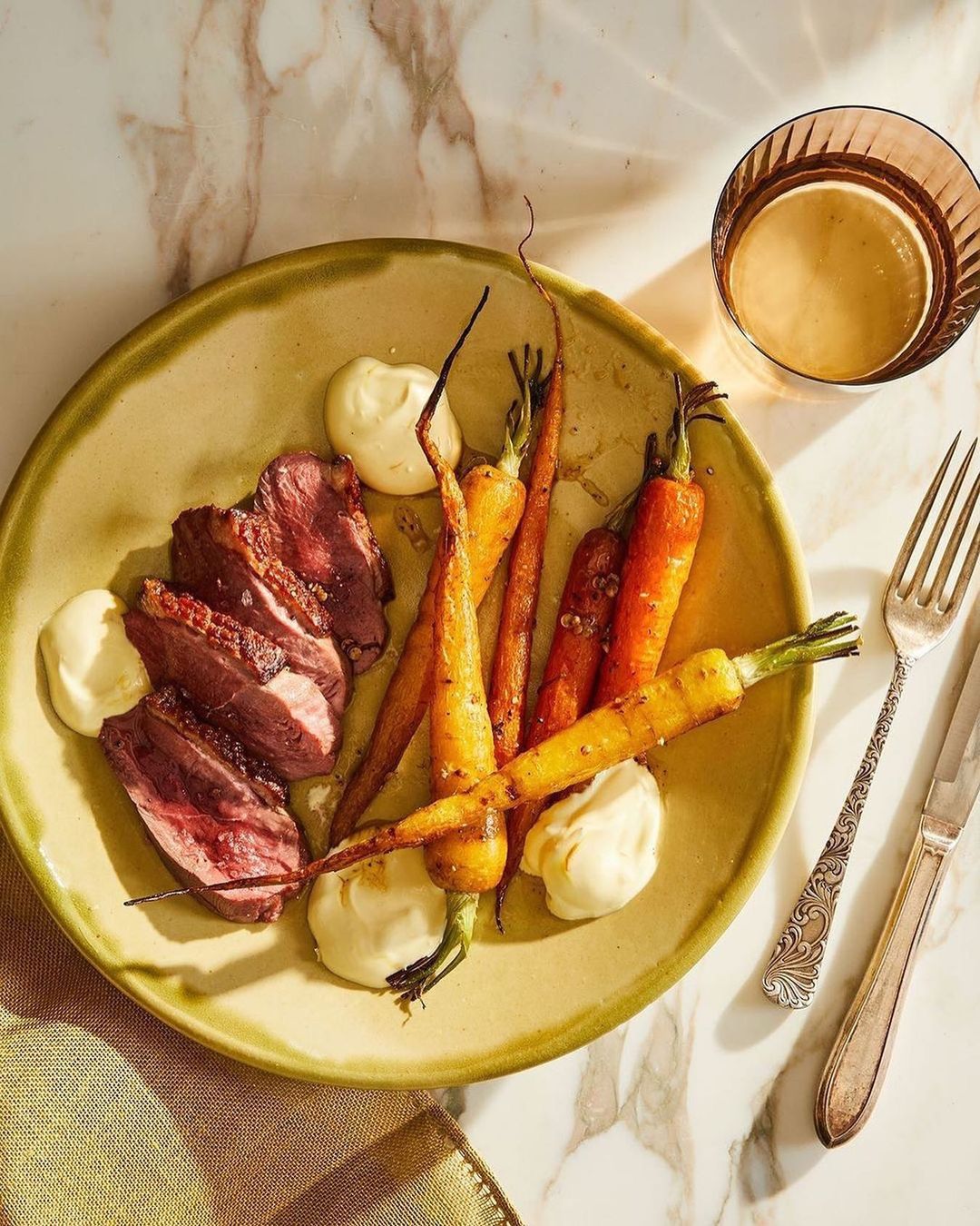 Overhead shot of a plate featuring sliced duck breast, roasted carrots, and dollops of creamy sauce, with silverware and drink nearby.