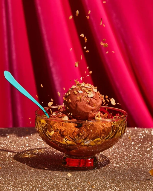 Chocolate ice cream with nut toppings in a decorative glass bowl on a glittery surface against a draped pink fabric backdrop.