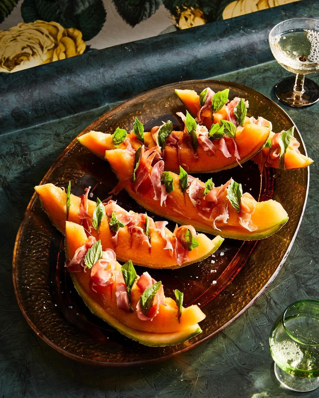 Overhead shot of cantaloupe slices topped with prosciutto and mint on a bronze platter, with champagne and floral wallpaper.