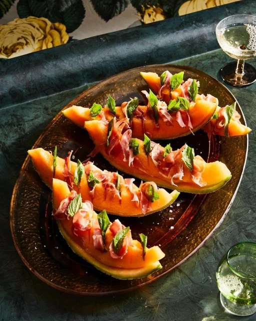 Overhead shot of cantaloupe slices topped with prosciutto and mint on a bronze platter, with champagne and floral wallpaper.
