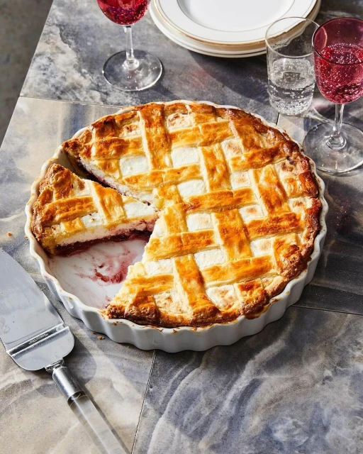 Overhead shot of a lattice-topped pie on a marble surface, accompanied by glasses of red liquid and a silver serving utensil.