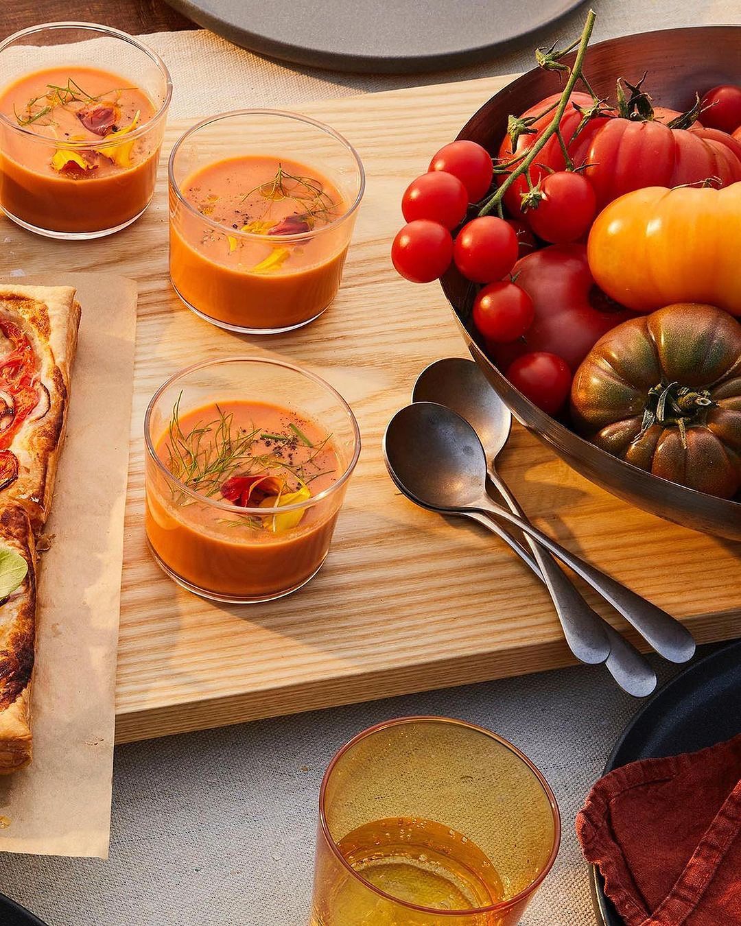 Overhead shot of glasses of orange soup garnished with herbs, accompanied by tomatoes and a tomato tart, set on a wooden surface.