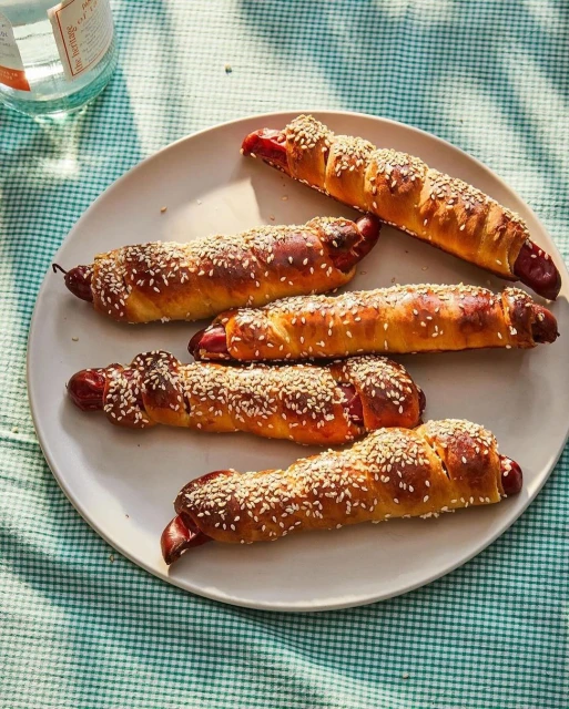 Overhead shot of a plate with five sausage rolls topped with sesame seeds, set on a blue and white gingham tablecloth next to a glass bottle.