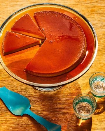 Overhead shot of a sliced flan dessert on a glass stand, with two drinking glasses and a serving utensil on a wooden surface.