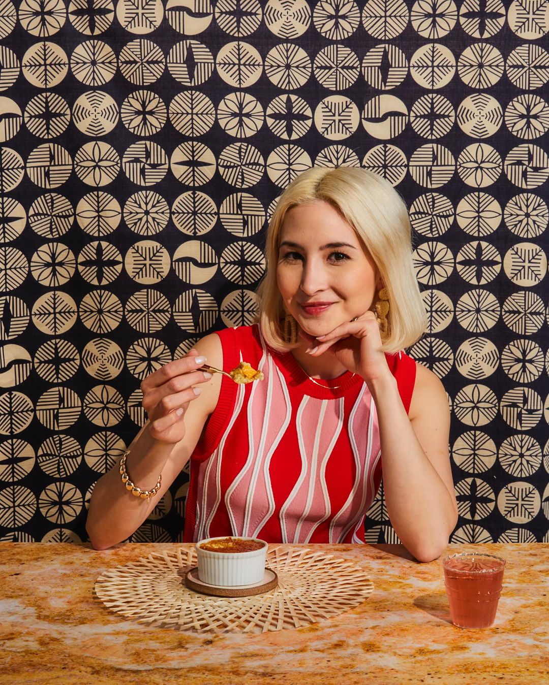 Woman poses at table set with crème brûlée and a beverage, wearing a patterned top with blonde hair and gold jewelry against patterned background.