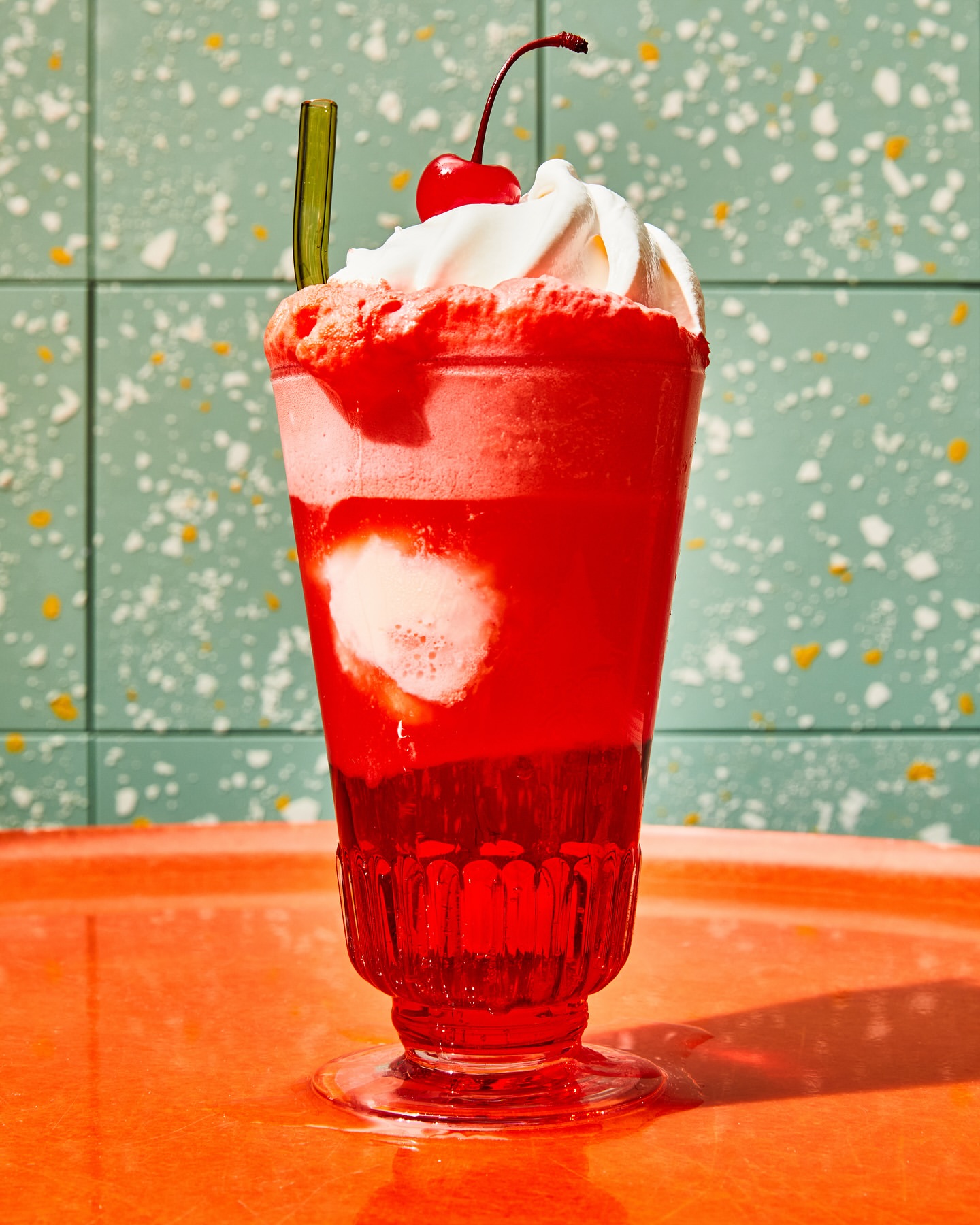 Tall dessert glass filled with a layered red beverage topped with whipped cream and a cherry, standing on an orange surface.