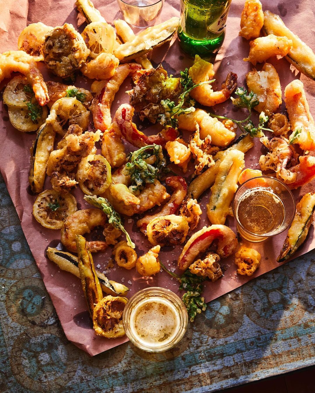 Overhead shot of tempura battered vegetables and shrimp served with dipping sauces and beer on a pink paper-lined rustic table.
