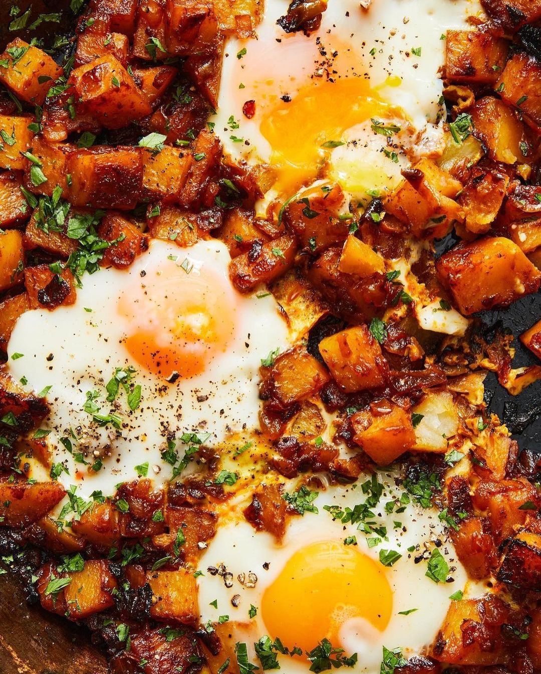 Overhead view of cooked potatoes with three fried eggs, seasoned with herbs and spices. A savory and colorful food presentation.