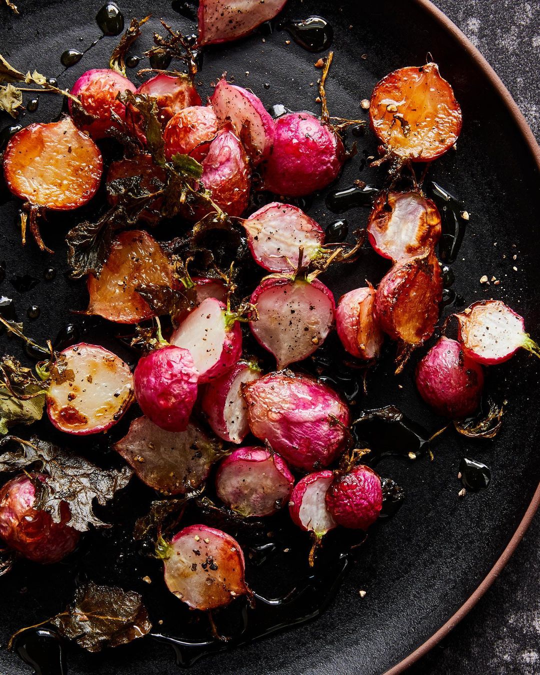 Overhead shot of roasted radishes on a black plate with dark sauce and leaves, showcasing vibrant colors and textures, food photography.