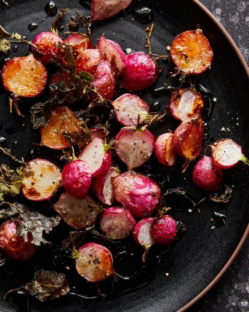 Overhead shot of roasted radishes on a black plate with dark sauce and leaves, showcasing vibrant colors and textures, food photography.