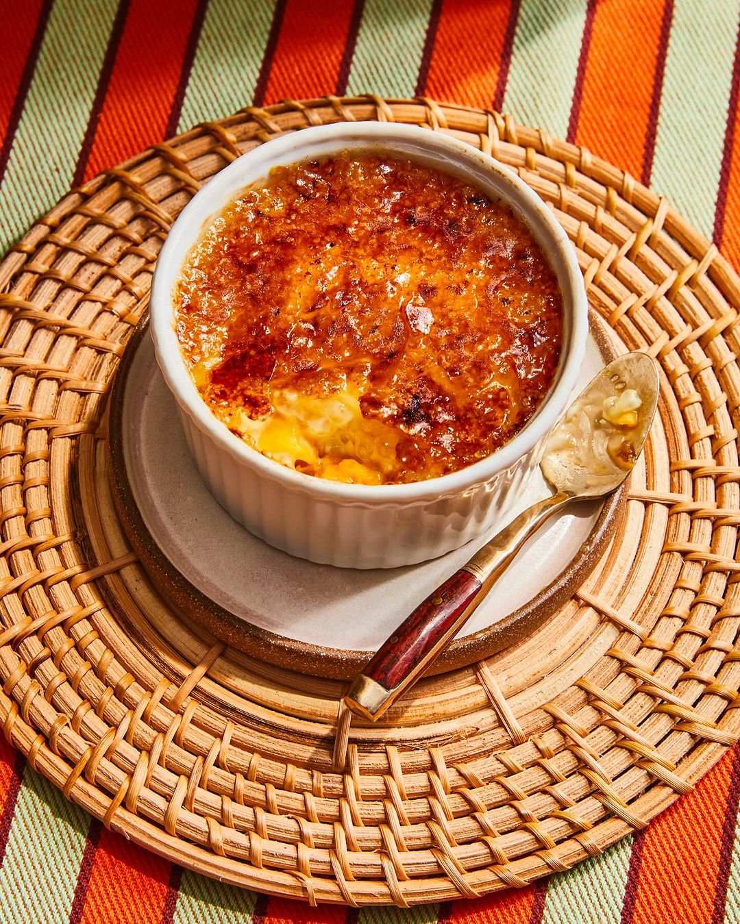 Creme brulee in white ramekin on a woven mat with striped fabric backdrop and a spoon, highlighting the dessert's caramelized topping.
