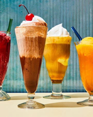 Four colorful ice cream floats in tall clear glasses with straws and whipped cream are arranged on a light yellow table.