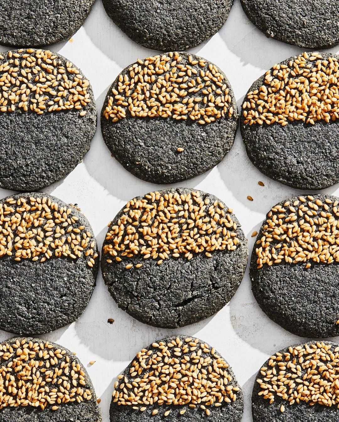 Overhead shot of black sesame cookies halved with sesame seeds on a white surface, food photography, baking, dessert, snack.