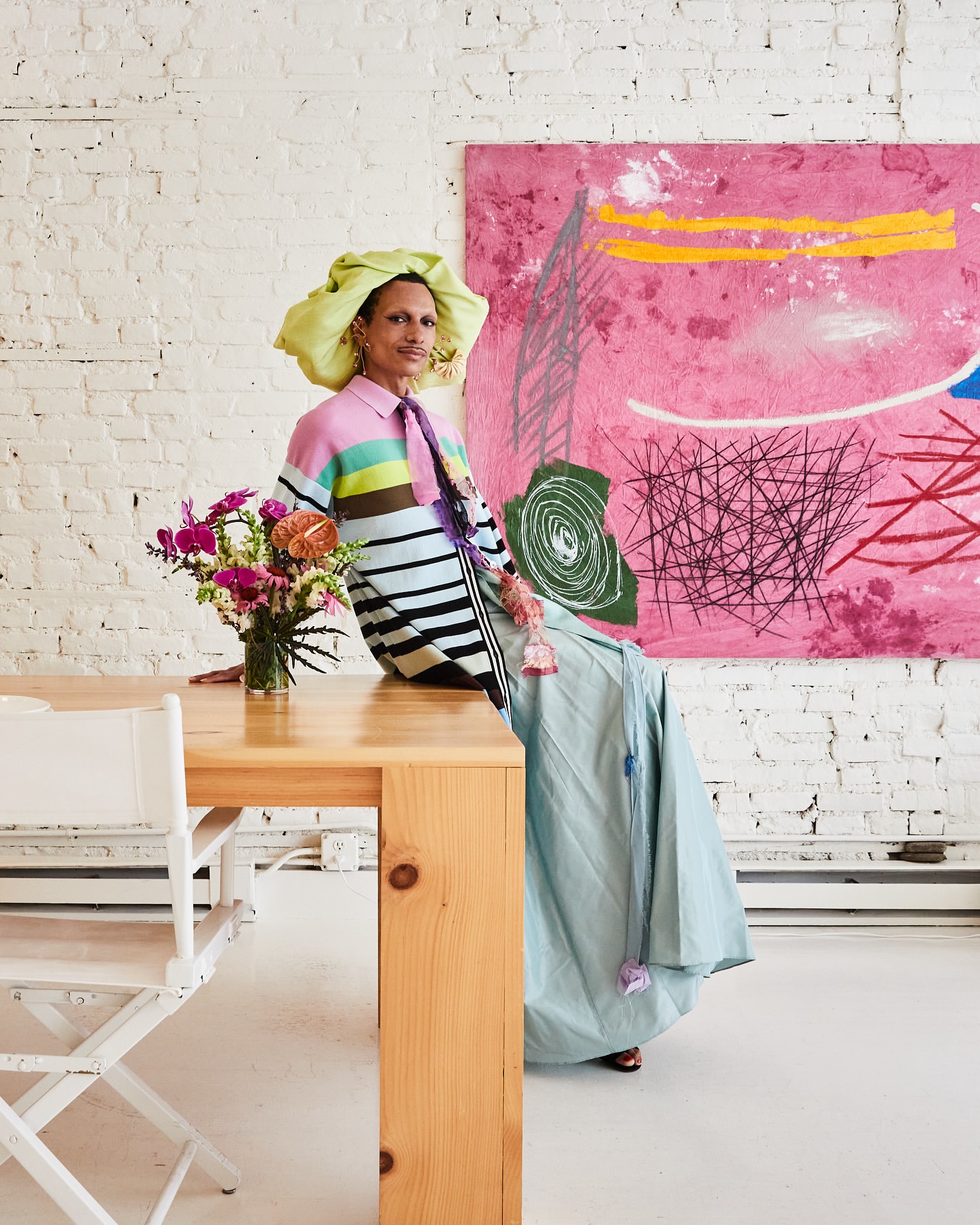 Person sits on table in bright fashion ensemble and large hat, beside a vase of flowers, in front of an abstract art painting on brick wall.