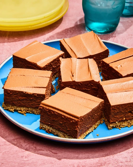 Square pieces of chocolate dessert with frosting on a blue plate, presented on a pink surface near a clear glass and a yellow plate.