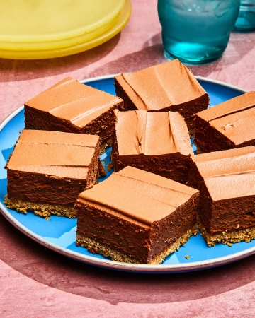 Square pieces of chocolate dessert with frosting on a blue plate, presented on a pink surface near a clear glass and a yellow plate.