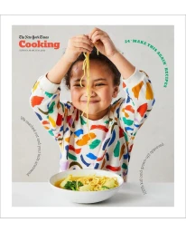 Child smiling while holding up noodles from a bowl, wearing a colorful patterned sweatshirt in a studio setting.