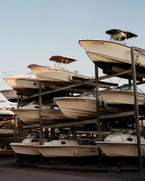 Multiple white motorboats stored on a multi-tiered metal rack structure outdoors under a clear pale sky.
