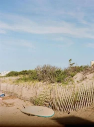 A surfboard resting on sandy ground near a wooden slat fence against sand dunes and a blue sky.