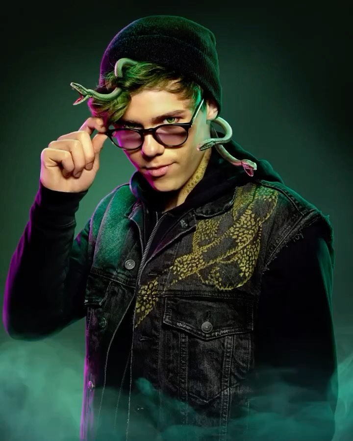 Portrait of a young adult with green hair, wearing a beanie and glasses, adorned with snakes, against a green studio backdrop.