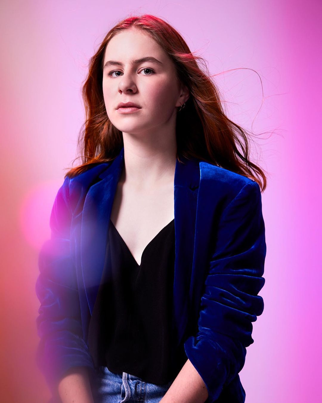 Portrait of a young adult woman with auburn hair wearing a blue velvet blazer and black top, against a pink and purple background.