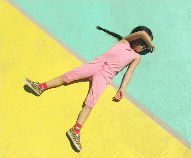 Child in pink jumpsuit lies diagonally on yellow and turquoise surface, shielding eyes from sun. Black braided hair, patterned shoes.