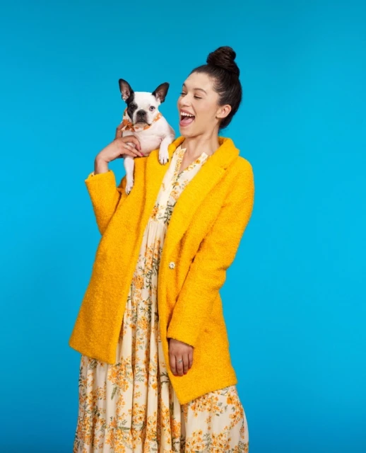 Cheerful young woman in floral dress and yellow coat poses with a Boston terrier puppy in a bright blue studio environment.