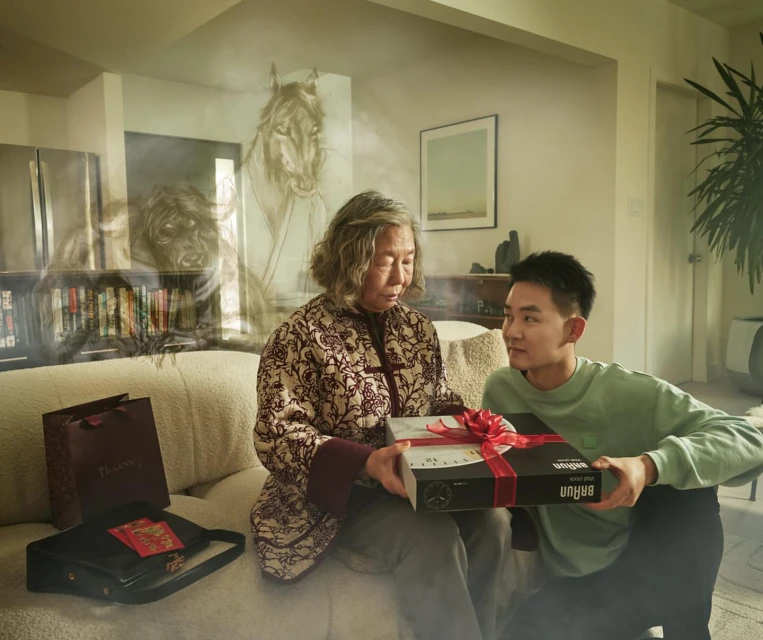 Elderly woman receiving a gift from a younger man indoors, with a painting visible in the background, creating a warm, generational moment.