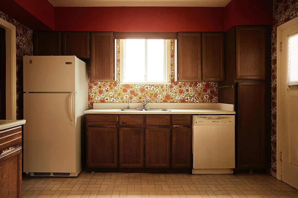 A retro kitchen interior featuring dark wood cabinetry, a white refrigerator, and a floral wallpaper backsplash under a window.